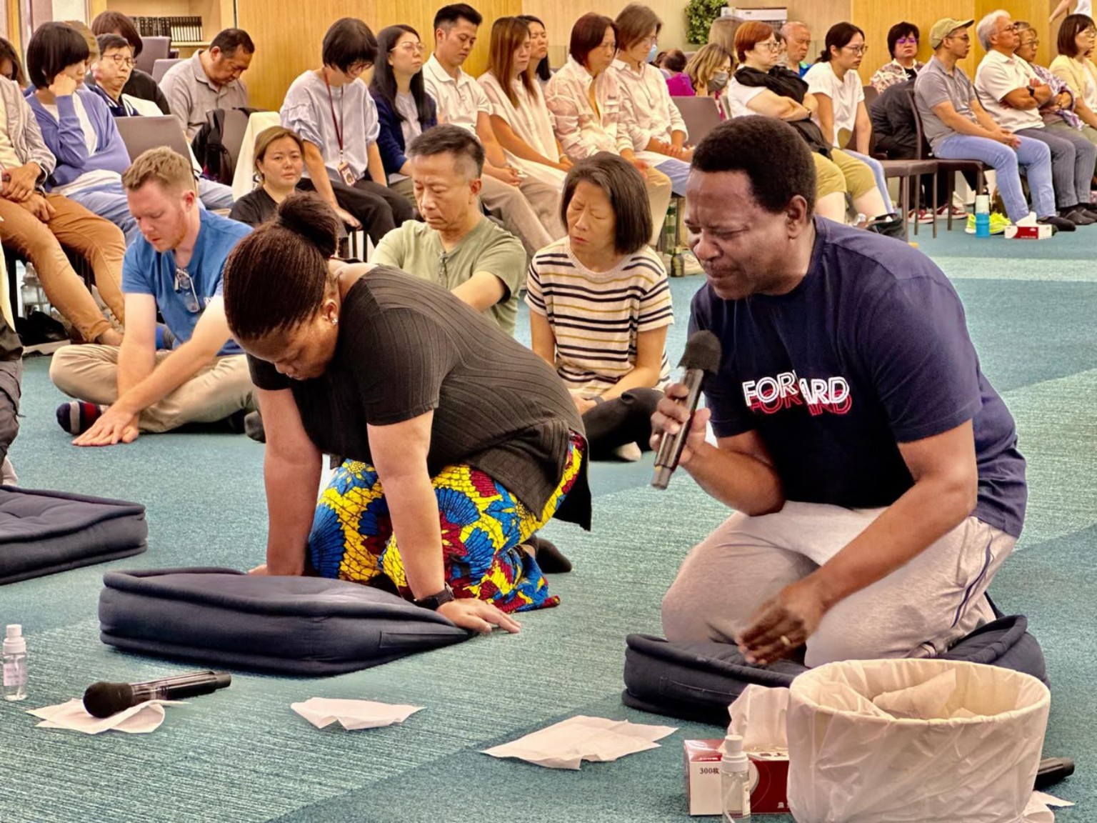Two men kneeling in prayer in front of a seated congregation.