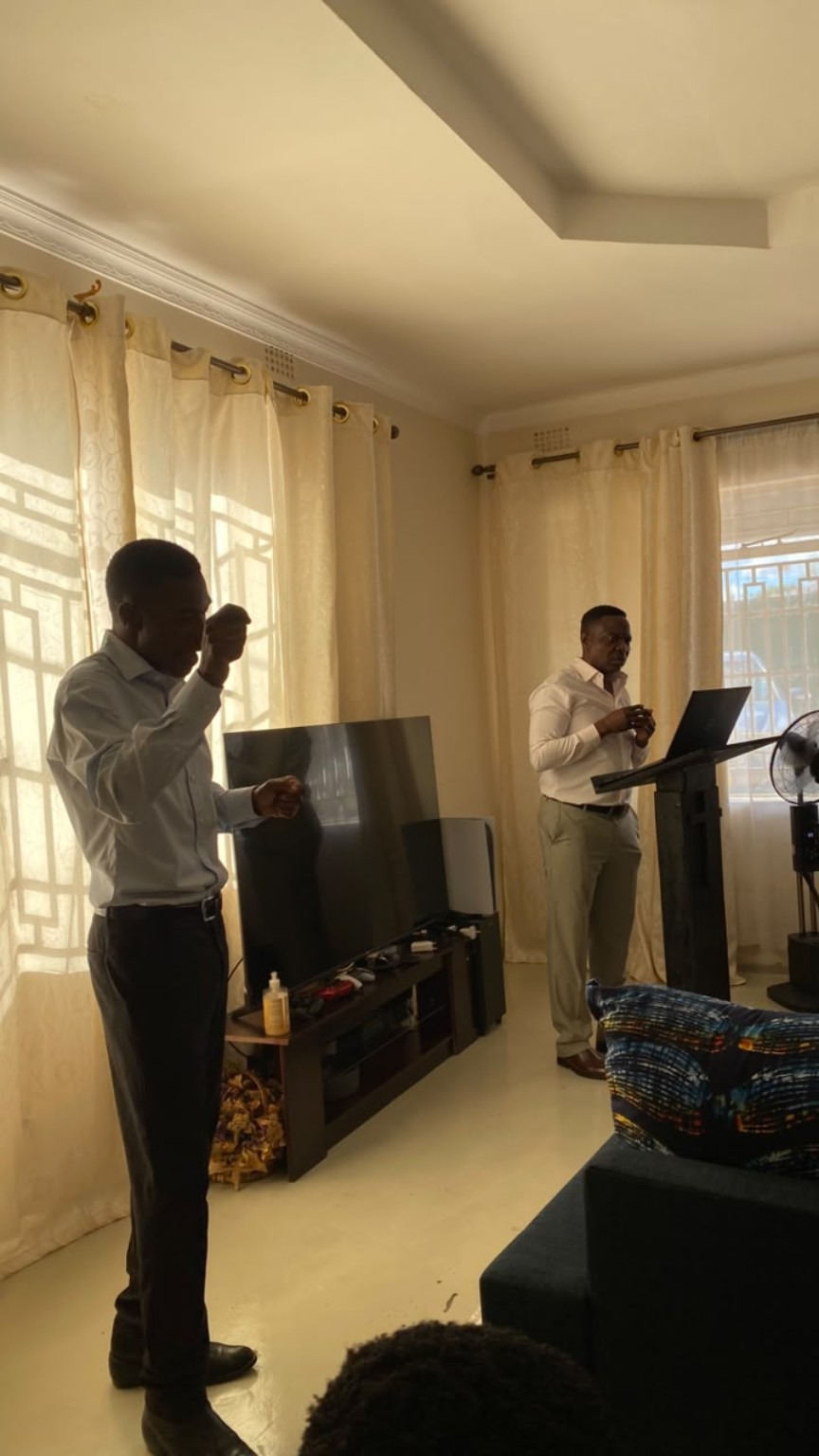 Two men standing during a gathering in a living room, one beside a lectern and laptop.