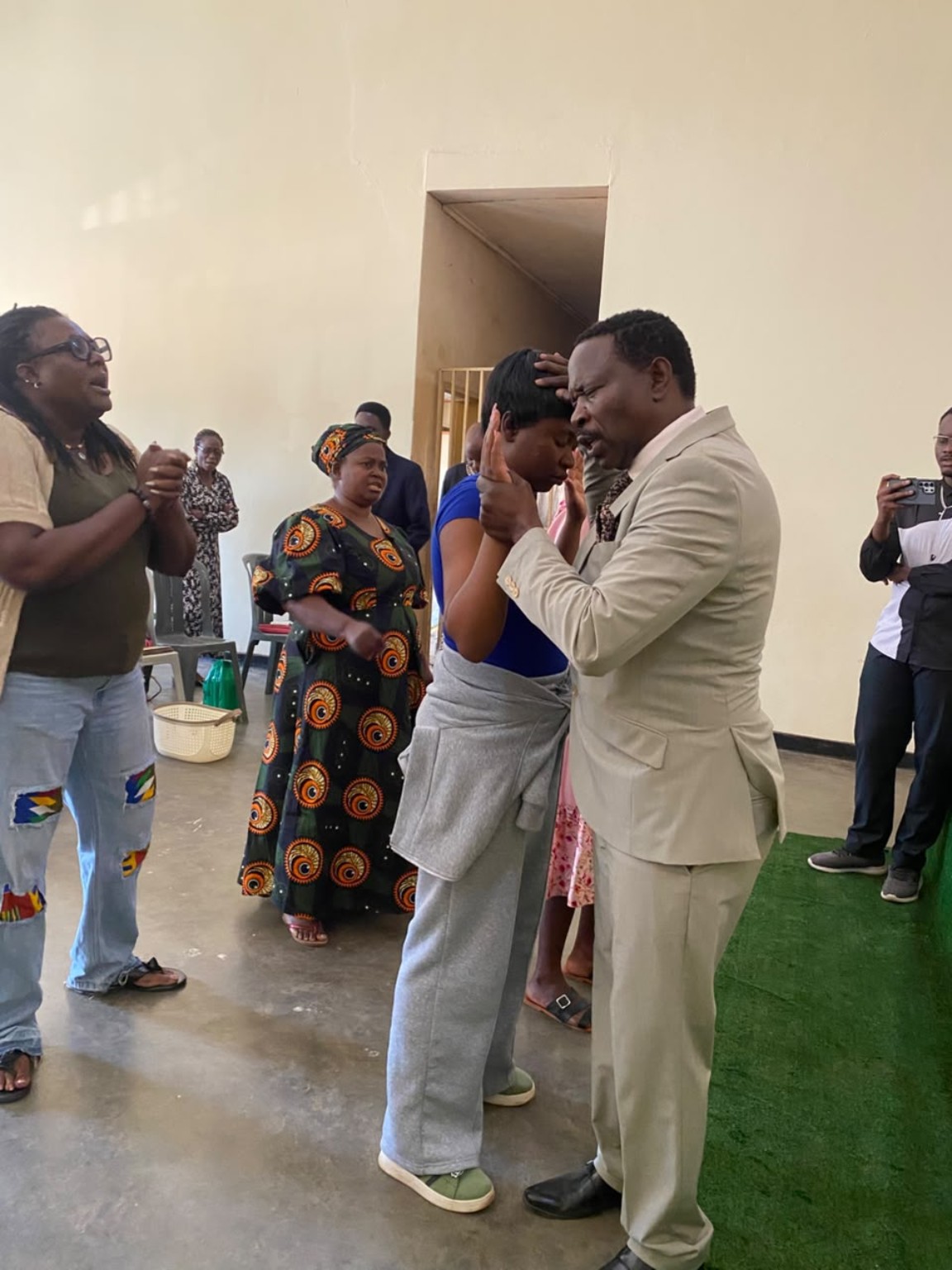 A ministry leader in a cream suit praying with a woman in blue as church members stand nearby.