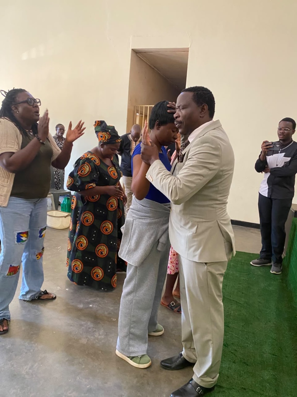 A ministry leader in a cream suit praying with a woman in blue as church members stand nearby.