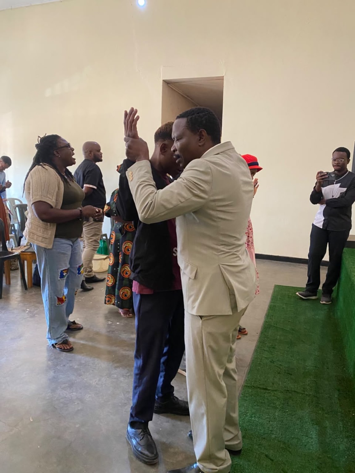 A ministry leader in a cream suit praying with a church member beside the green platform.
