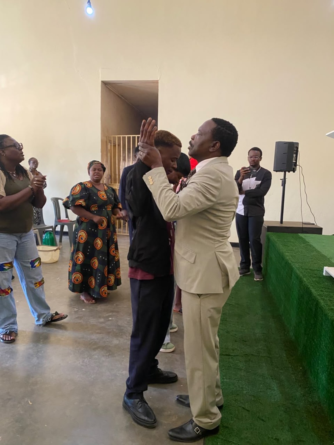 A ministry leader in a cream suit praying with a church member beside the green platform.