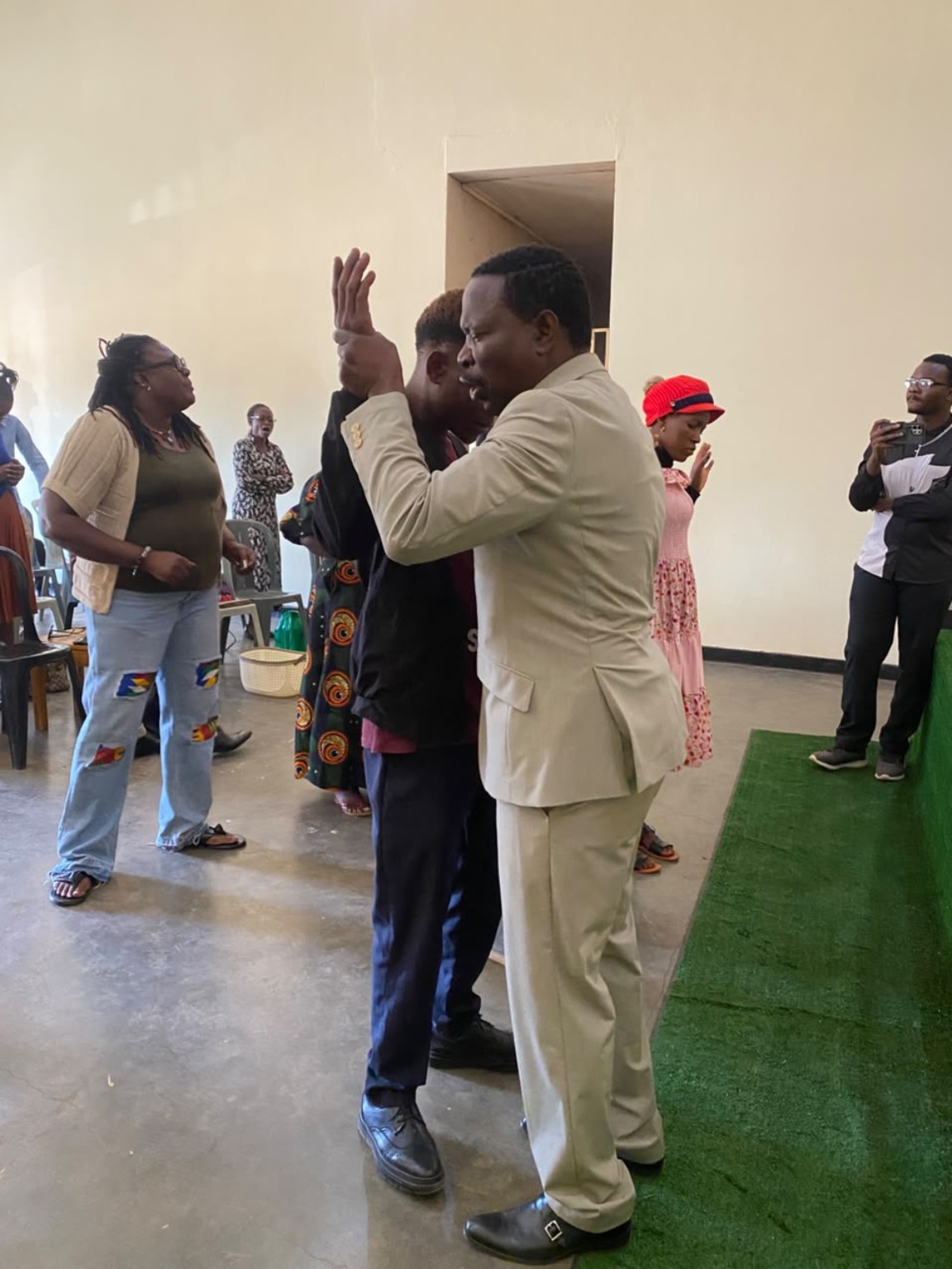 A ministry leader in a cream suit praying with a church member beside the green platform.