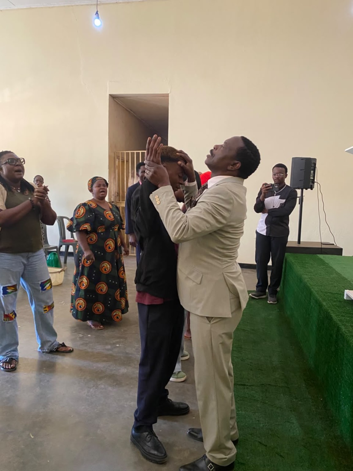 A ministry leader in a cream suit praying with a church member beside the green platform.