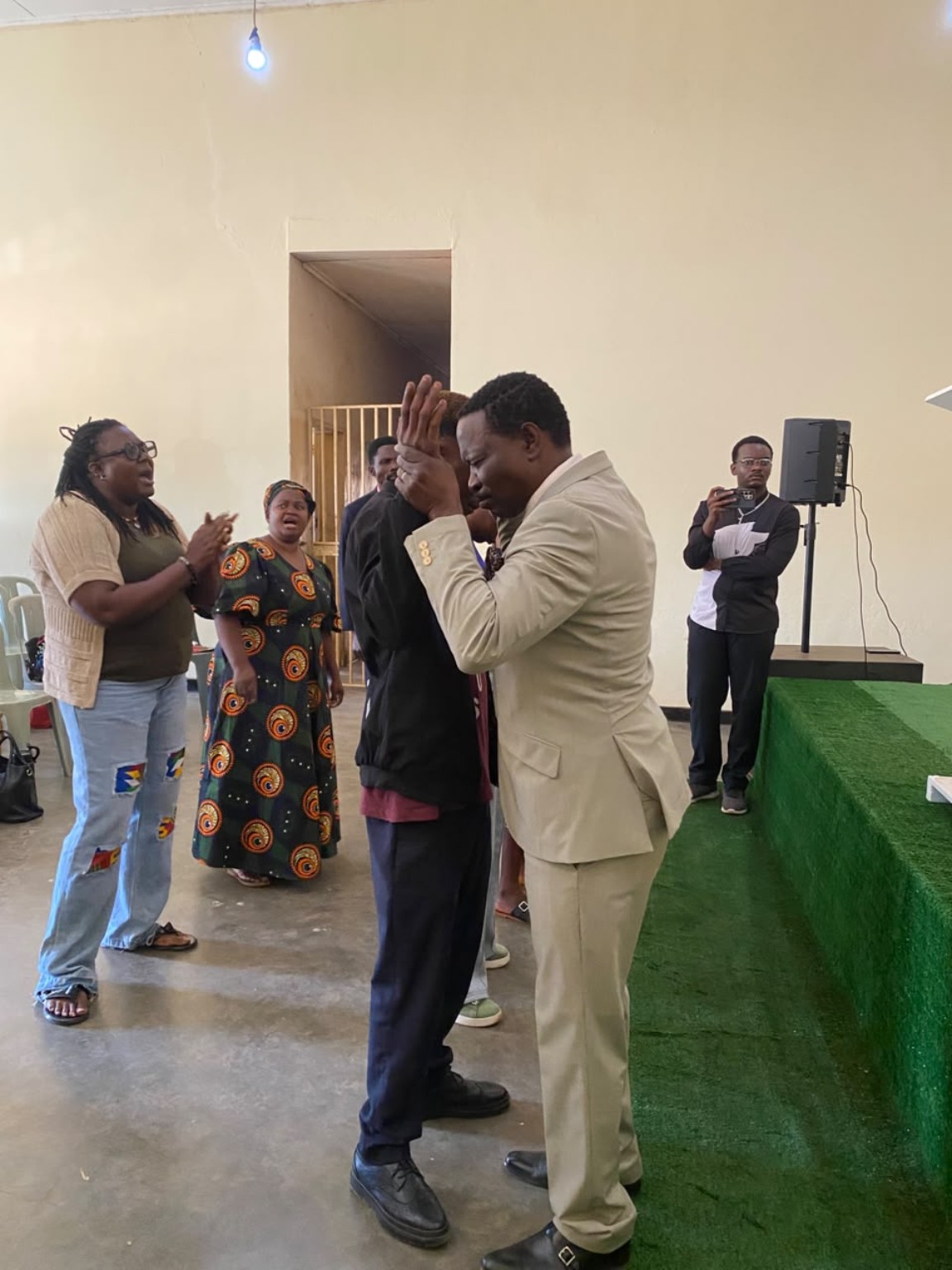 A ministry leader in a cream suit praying with a church member beside the green platform.