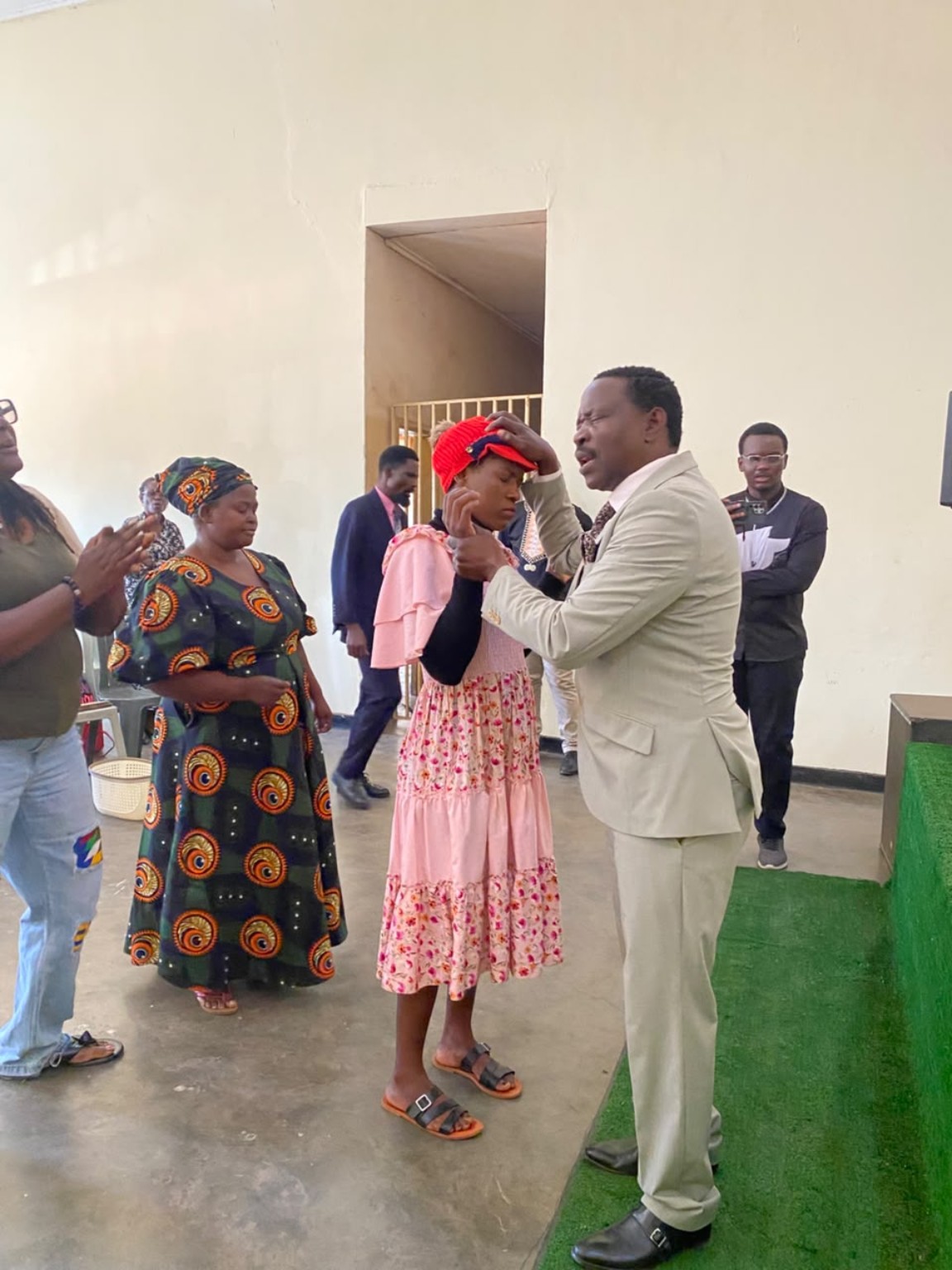 A ministry leader in a cream suit praying with a woman wearing a red hat and pink dress.