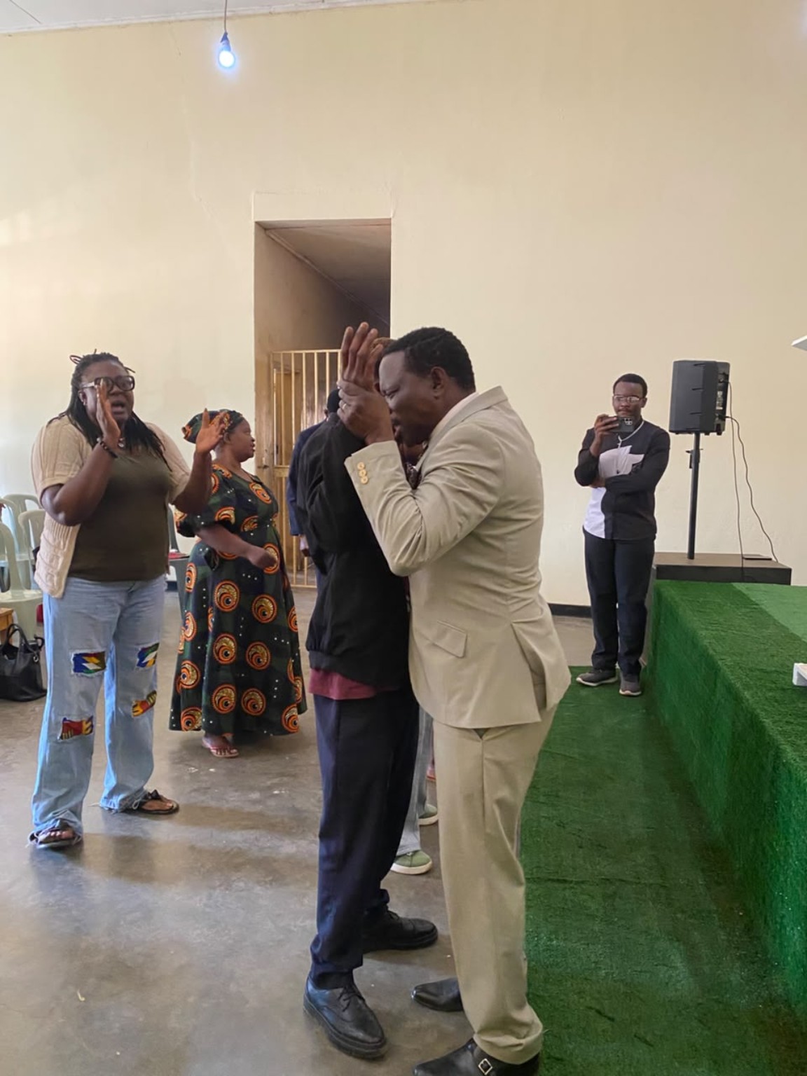 A ministry leader in a cream suit praying with a church member beside the green platform.
